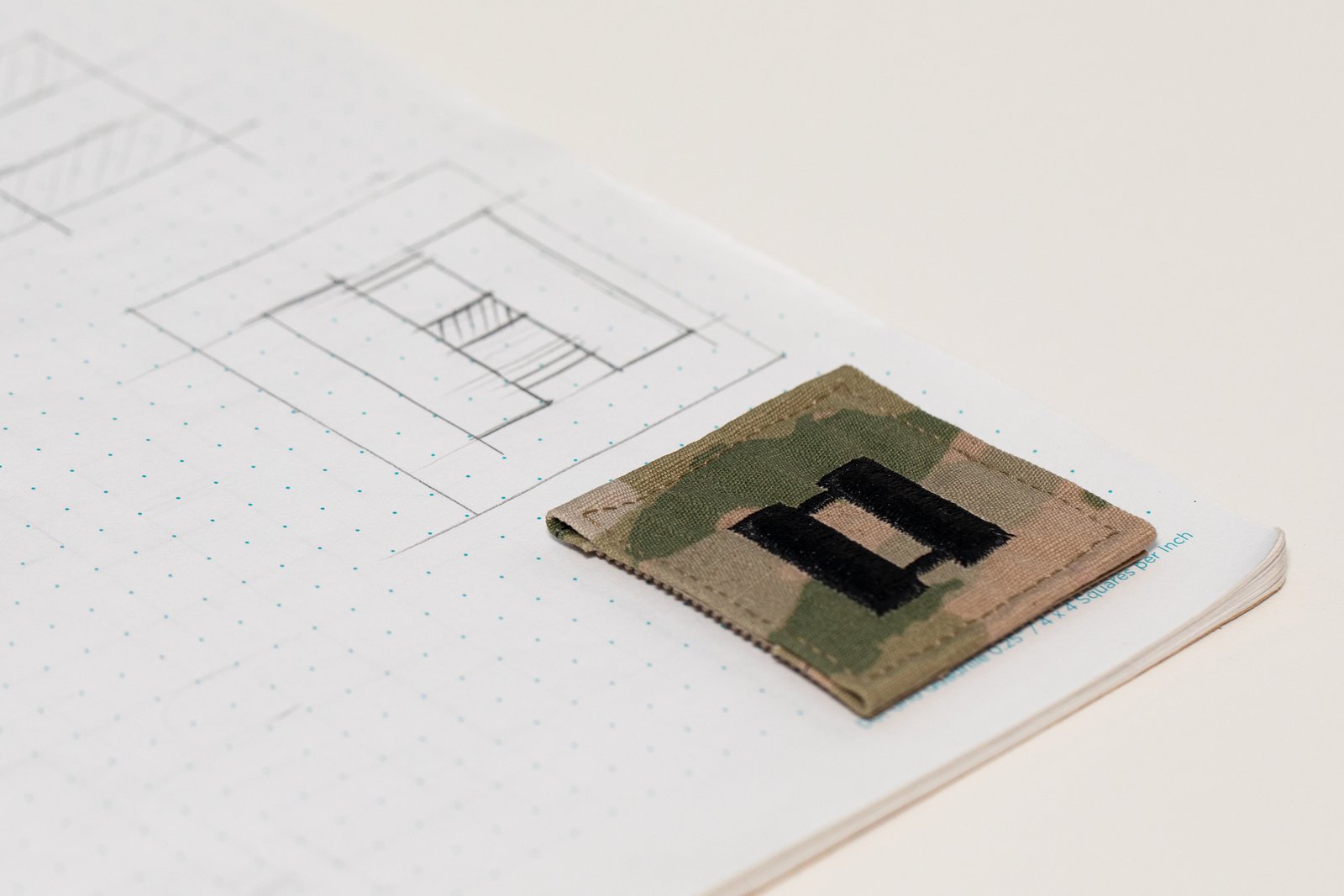 Sketchbook page with DHC logo studies and a U.S. Army captain’s rank patch placed on the corner of the notebook.